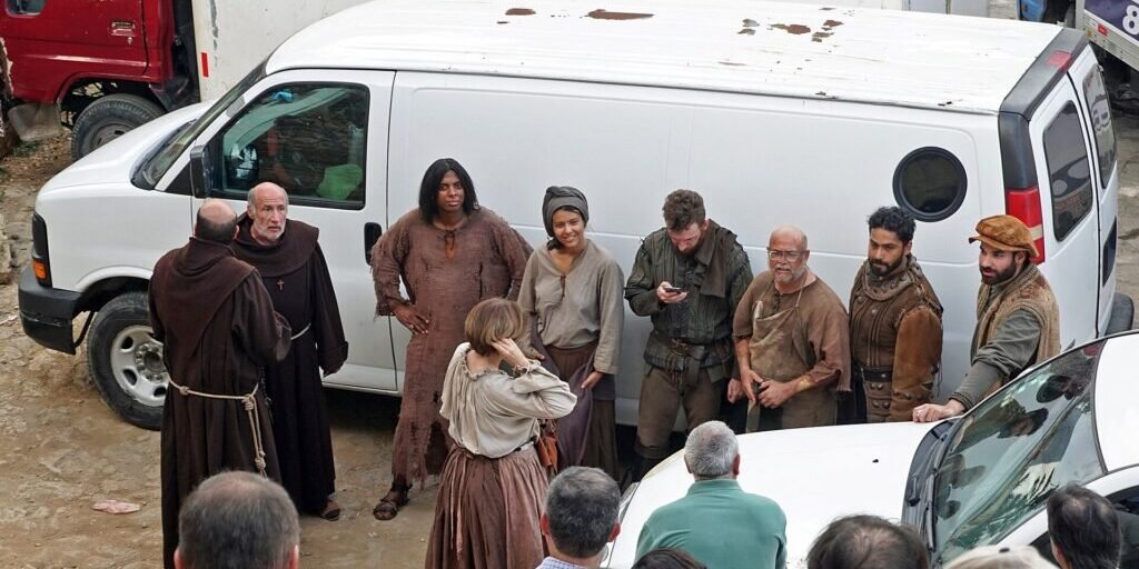 A group of actors in period costume stand together near production vans, taking a break on a film set