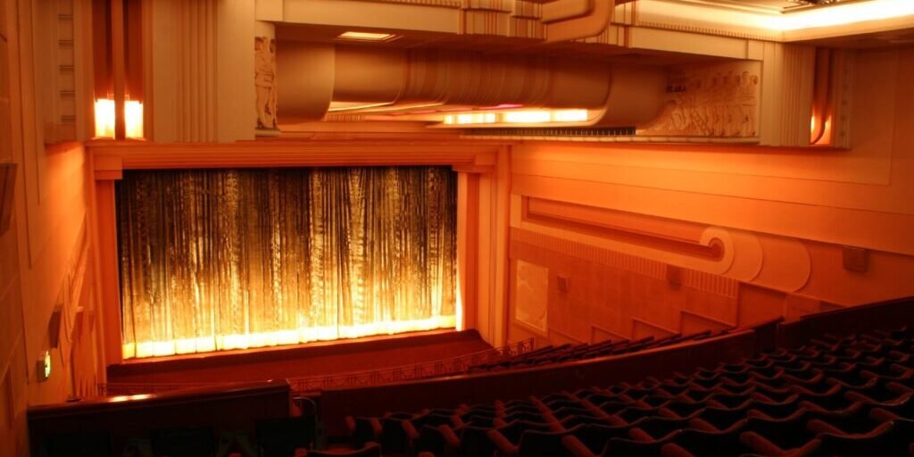 Ornate Art Deco cinema auditorium with illuminated curtained screen and rows of seats