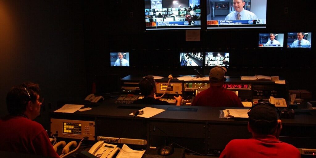 Producers at a console in a television broadcast control room with a wall of video monitors