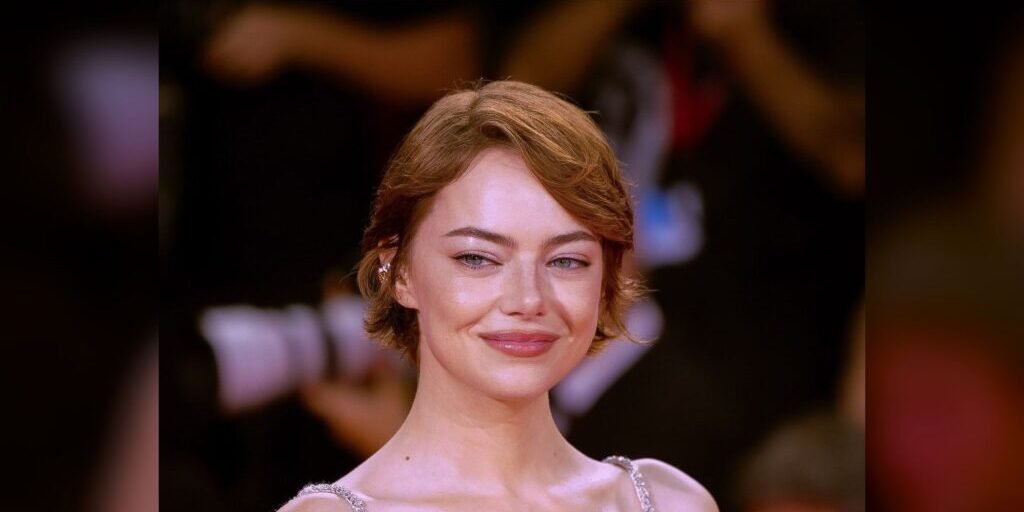 Emma Stone smiling on a red carpet in a silver beaded dress, with blurred photographers and a dark background behind her.