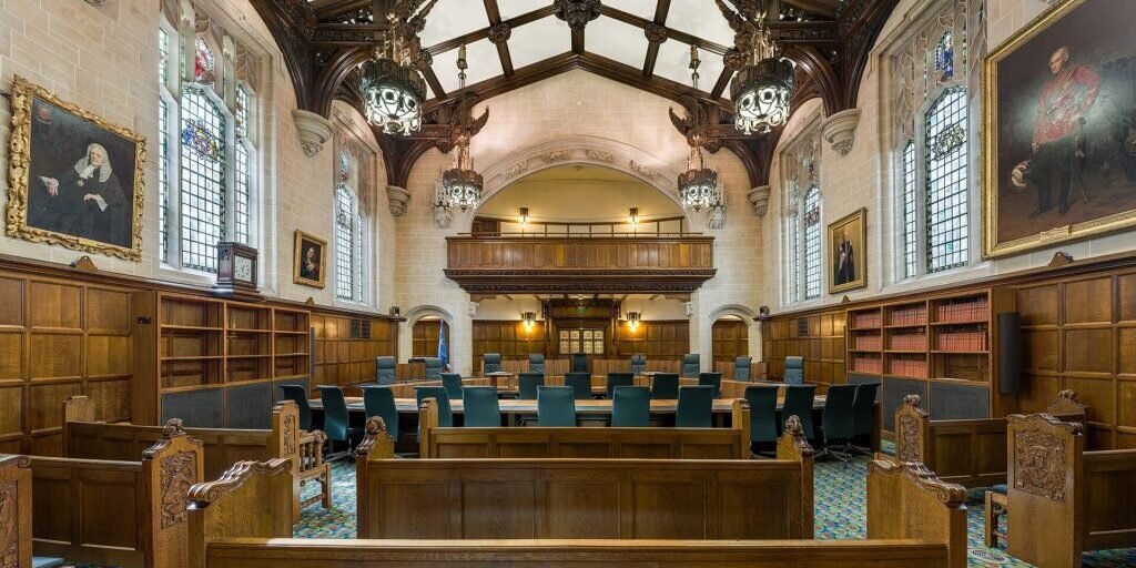 Empty wood-panelled courtroom with rows of benches and blue chairs under a vaulted timber ceiling.