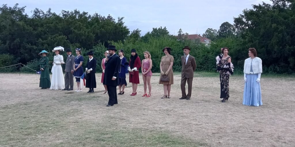 People in period costume with hats and long dresses stand in a line on a grassy outdoor field in front of trees.