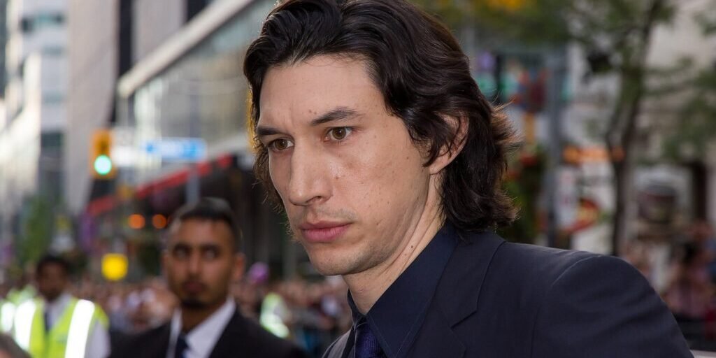 Adam Driver in a dark suit and tie outdoors, looking to the side, with a blurred crowd and street behind him