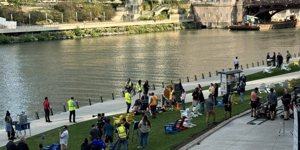 Television production set with crew members and equipment along a waterfront with city buildings